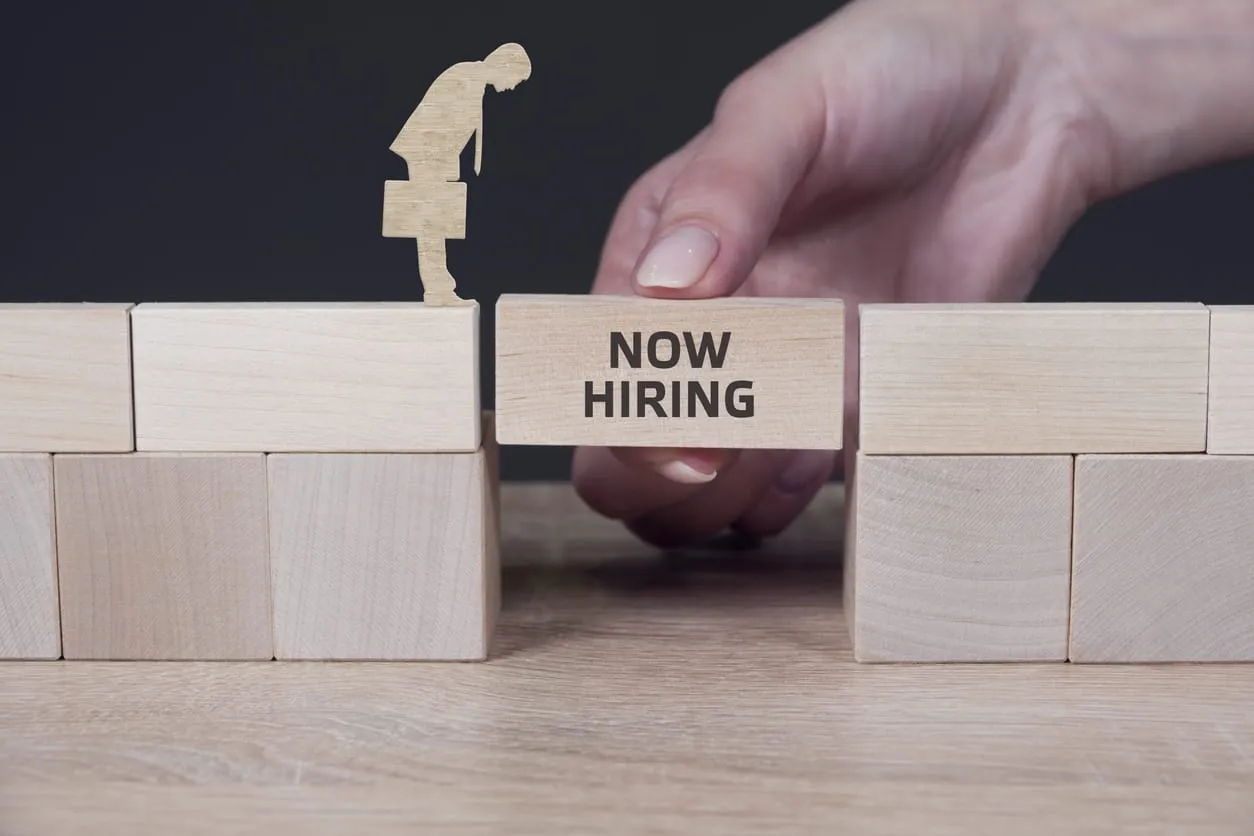 inverses-headhunting-karriere-weg Une main place un bloc de bois portant l’inscription « Now Hiring » entre deux espaces, créant ainsi un passage pour une figurine munie d’une mallette – une métaphore de l’headhunting inversé.